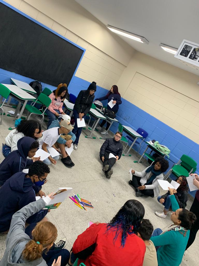A foto mostra um grupo de cerca de 15 jovens em uma sala de aula; eles estão sentados no chão em um círculo, seguram folhas de papel nas mãos e conversam entre si
